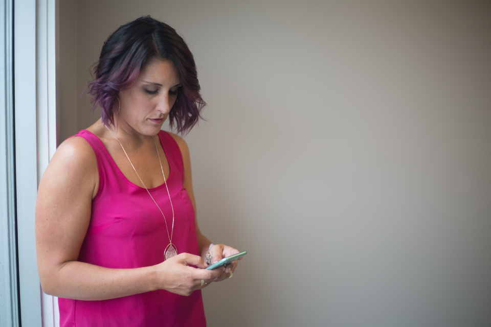 Mulher olhando para o celular enquanto digita uma mensagem, em pé perto de uma janela