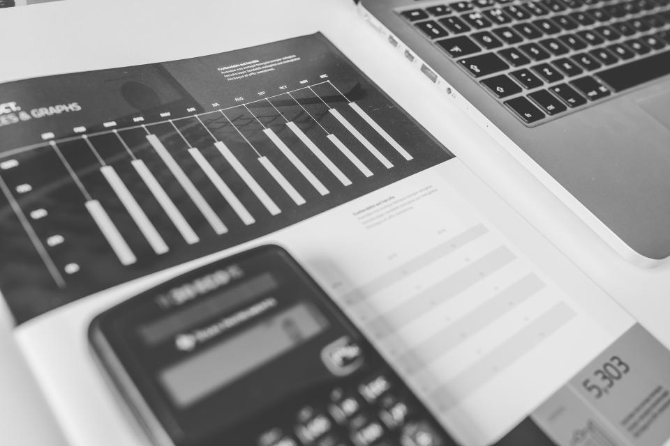 Calculadora sobre uma revista com gráficos, ao lado de um notebook, em preto e branco