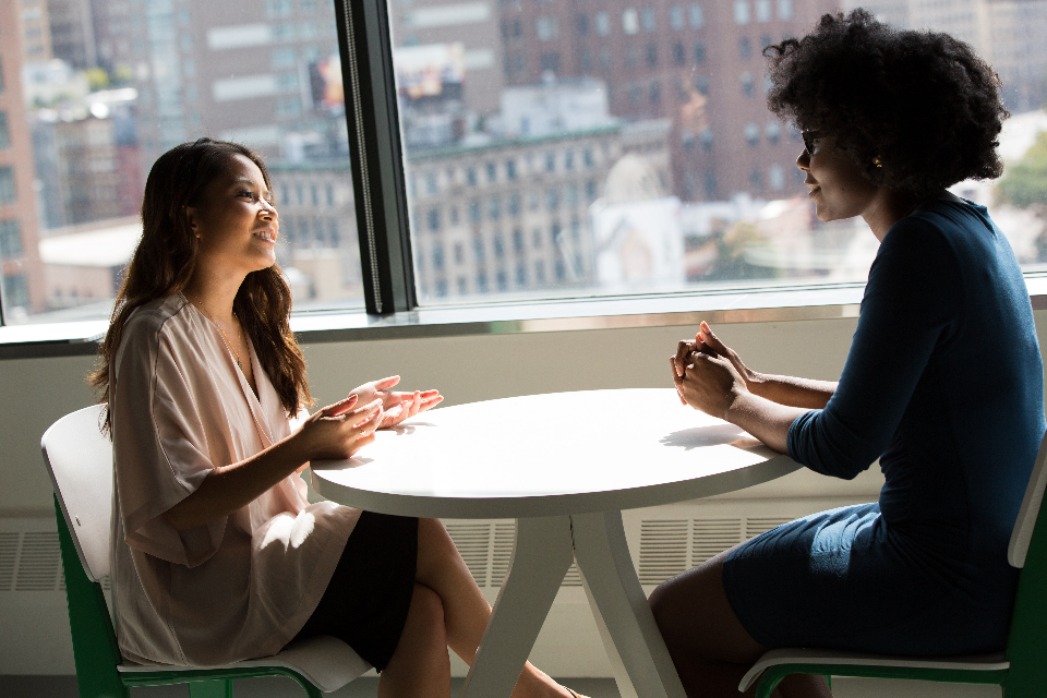 Duas mulheres conversando sentadas em uma mesa redonda, em um escritório com vista para prédios
