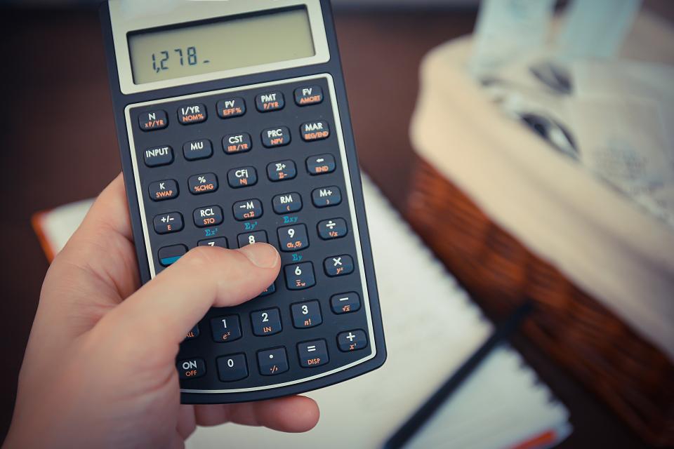 Mão segurando uma calculadora financeira com o número 1.278 no visor, sobre uma mesa com anotações ao fundo