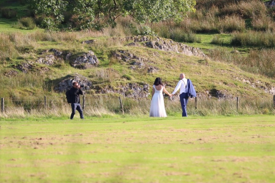 Fotógrafo de roupa escura fotografando um casal de noivos de mãos dadas num campo aberto, com colina e pedras ao fundo
