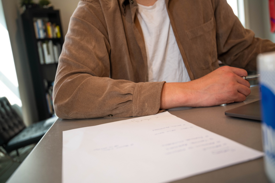 Pessoa revisando um documento impresso sobre a mesa, ao lado de um notebook fechado