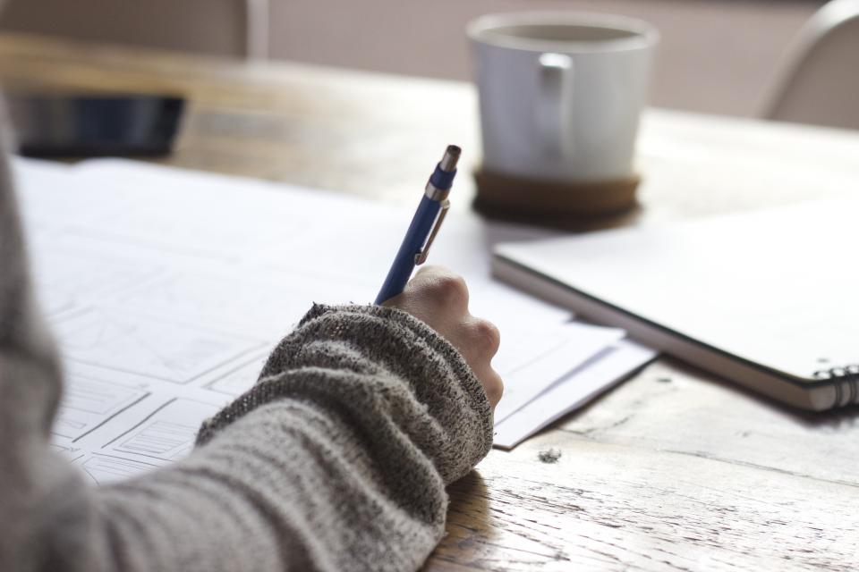 Pessoa escrevendo anotações num caderno sobre a mesa, com xícara de café ao lado