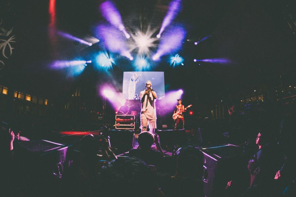 Show visto do meio do público, com artista no palco e luzes coloridas de refletor ao fundo