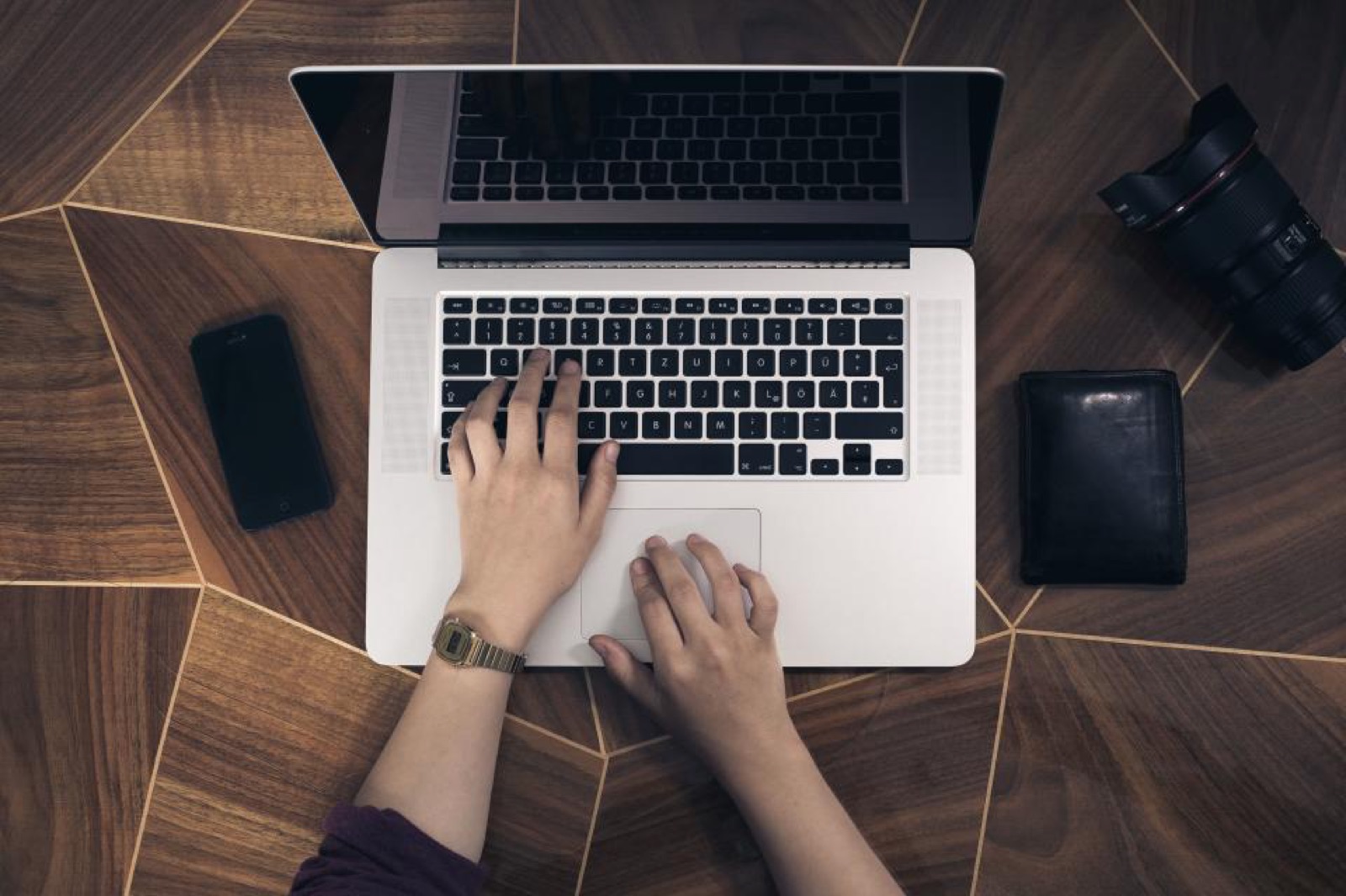Vista de cima de um notebook aberto sobre uma mesa de madeira com desenho geométrico, duas mãos no teclado, um celular à esquerda, uma carteira preta à direita e a objetiva de uma câmera no canto superior direito