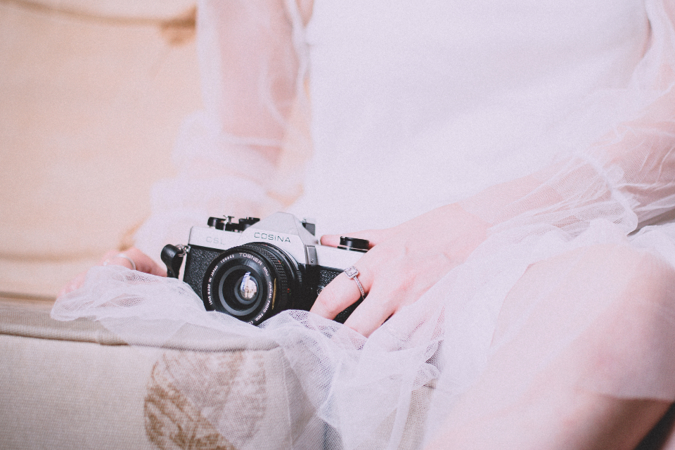 Mão de uma noiva com anel de noivado segurando uma câmera fotográfica vintage sobre o vestido de renda