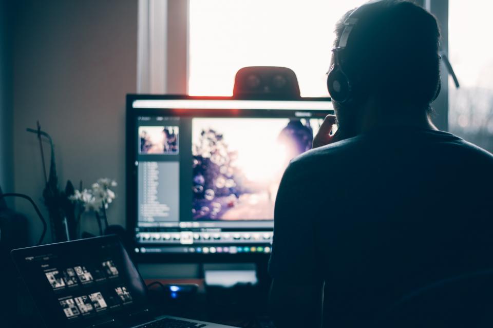 Editor de vídeo de costas, editando um vídeo num monitor, com um notebook ao lado