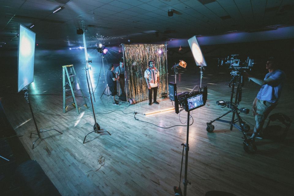 Set de gravação em estúdio com refletores, painel dourado ao fundo, câmera em tripé com rodinhas e equipe montando a cena