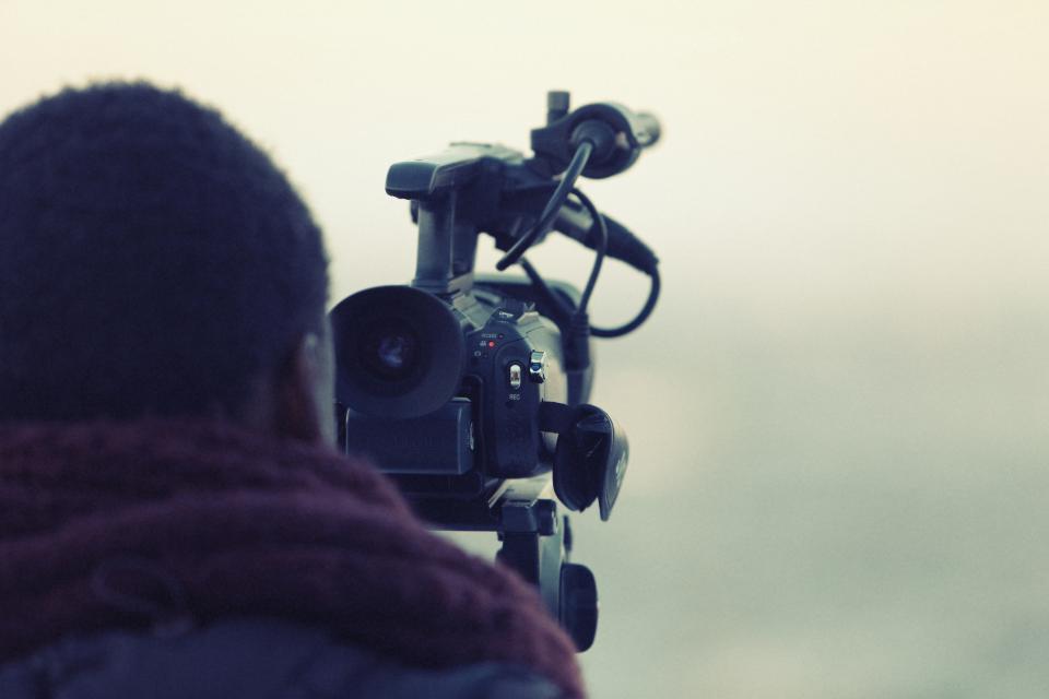 Videomaker filmando com câmera profissional e microfone acoplado, visto de costas