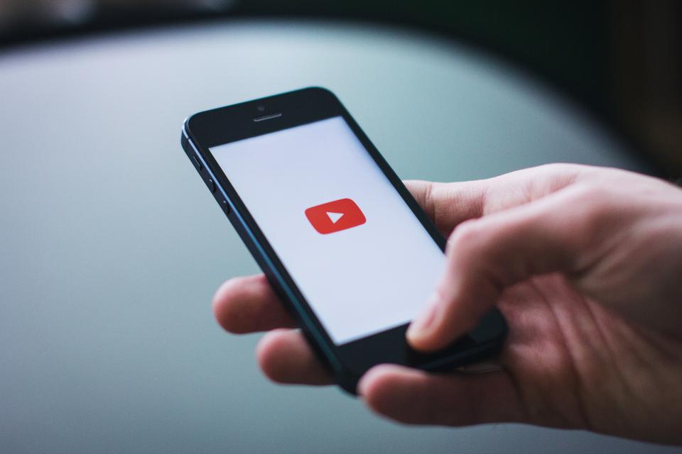 Mão segurando um iPhone com o aplicativo do YouTube abrindo na tela