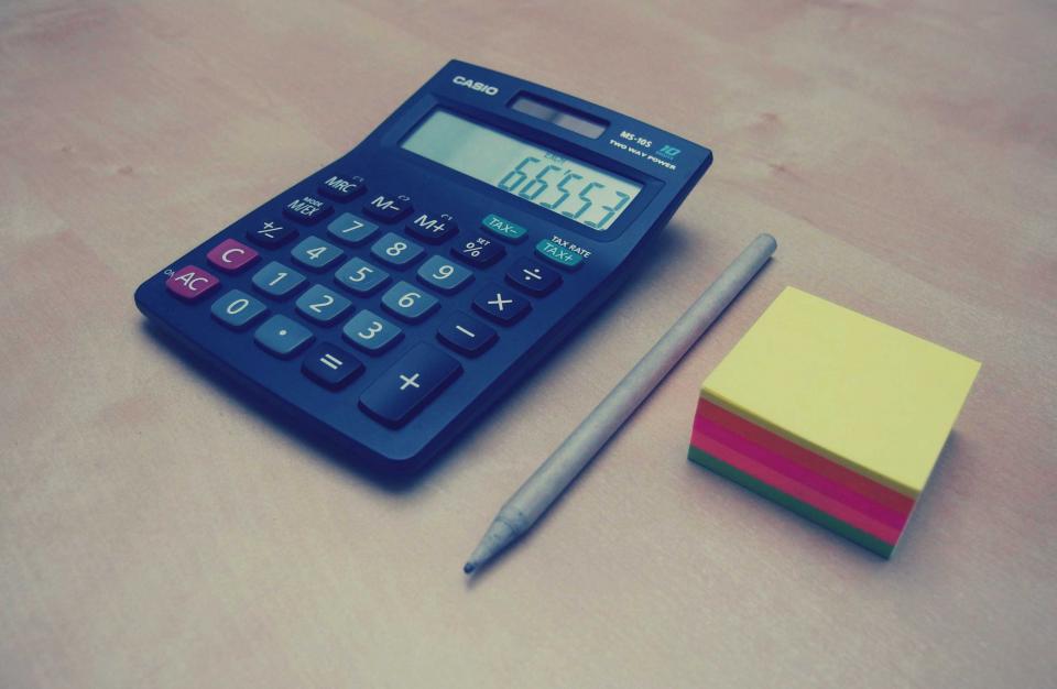 Calculadora com botão de taxa de imposto ao lado de um bloco de notas coloridas sobre uma mesa de madeira