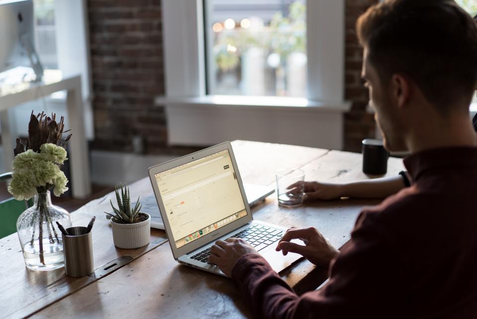 Homem trabalhando em um notebook sobre uma mesa de madeira em um escritório com plantas ao lado