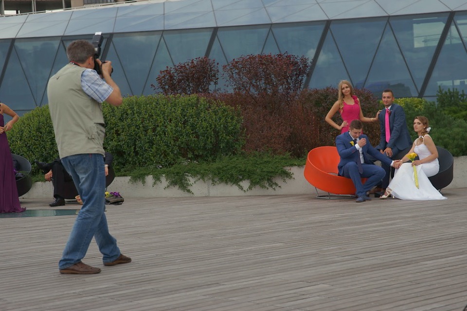 Fotógrafo de colete apontando a câmera para um casal de noivos e os padrinhos posando num deck, em frente a um prédio de vidro