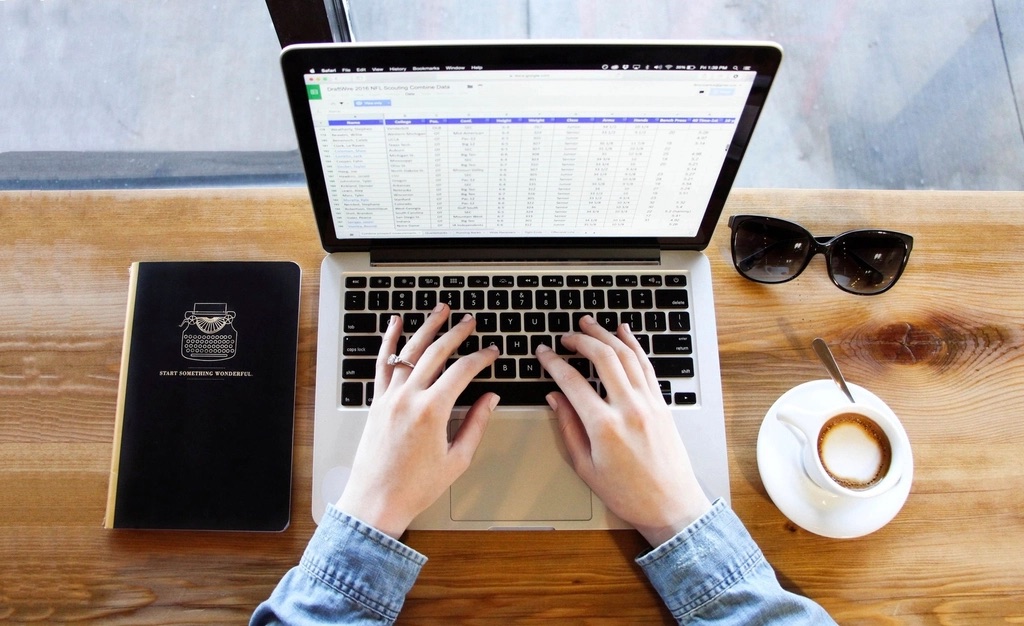 Mãos digitando num notebook com uma planilha aberta, sobre mesa de madeira com caderno, óculos de sol e café